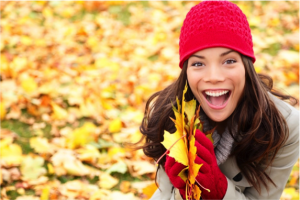 Joyful woman in a red hat and gloves and a beige coat holding autumn leaves
