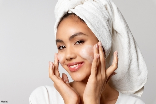 Calm woman with hair wrapped in a towel applying skincare cream to her cheeks and smiling