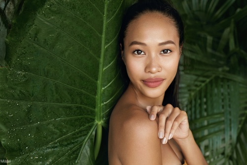 Tranquil, confident young woman with clear skin softly smiling in front of a background of tropical leaves